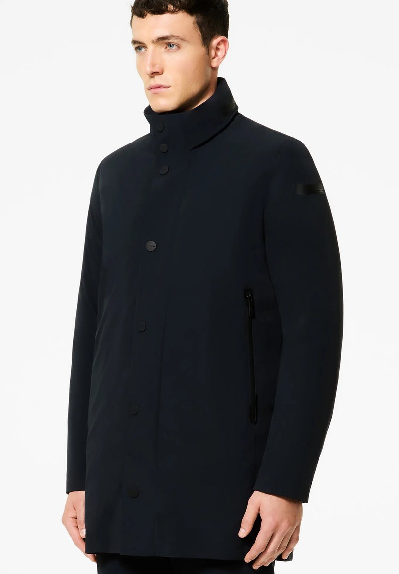 Uomo che indossa un cappotto nero a collo alto con chiusura a bottoni e tasca laterale con zip, rivolto leggermente verso sinistra su uno sfondo bianco uniforme.