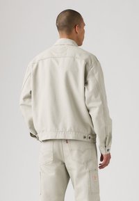 Chaqueta de mezclilla gris claro con un corte relajado, detalles de botones en los puños y sutiles acentos de costura. Llevada sobre pantalones a juego en gris claro.