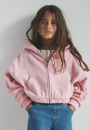 Mädchen mit langen braunen Haaren, trägt einen hellrosa Zip-up-Hoodie und blaue Jeans, steht mit den Händen in den Taschen vor einer schlichten weißen Wand.