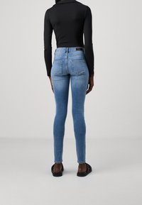 Person med ljusblå tajta jeans, svart långärmad tröja och svarta sandaler, står vänd bort från kameran mot en enkel grå bakgrund.