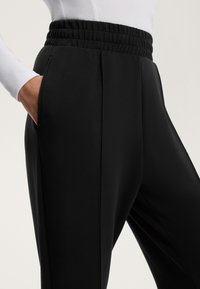 Čierne nohavice vyrobené z mäkkého materiálu s elastickým pásom, s bočnými vreckami a úzkym strihom, zdôrazňujúcim hladkú textúru.