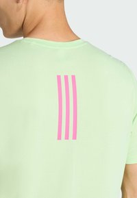 Camisa deportiva verde claro con una textura suave, mangas cortas y tres franjas verticales rosas en la parte superior de la espalda.