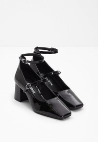 Scarpe con tacco a blocco in pelle verniciata nera con punta quadrata, dotate di due cinturini regolabili alla caviglia e di una finitura liscia e lucida.