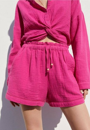 Femme portant une chemise à manches longues texturée rose vif nouée et un short à taille élastique avec cordon assorti, debout devant un fond clair.
