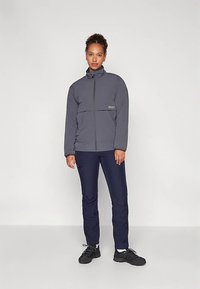 Giacca grigia con cerniera e collo alto, dotata di tasca sul petto. Abbinata a pantaloni aderenti blu navy e scarpe da ginnastica nere.