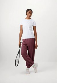 Λευκό εφαρμοστό μπλουζάκι συνδυασμένο με άνετο μπορντό παντελόνι φόρμας, κρατώντας ρακέτα τένις, με λευκά αθλητικά παπούτσια με διακριτικές ροζ λεπτομέρειες.