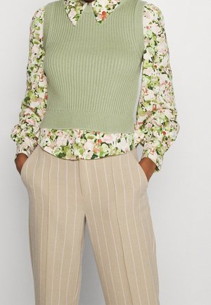 Person med en lys grøn ribbet ærmeløs sweater over en blomsterprintet bluse og beige bukser med striber, hænderne i lommerne.