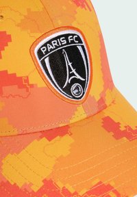 Kappe aus glattem Stoff in Orange und Gelb mit einem pixelierten Muster. Verfügt über ein schwarzes, rundes Logo mit "PARIS FC" und dem Eiffelturm.
