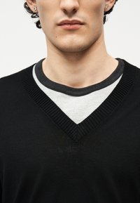 Νεαρός ενήλικας που φοράει μαύρο πουλόβερ τύπου V-neck πάνω από ανοιχτό γκρι μπλουζάκι με στρογγυλή λαιμόκοψη, με μικρά σκουλαρίκια ορατά και στα δύο αυτιά.