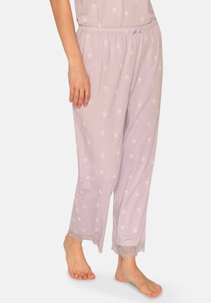 Frau trägt lockere, hellviolette Schlafanzughosen mit weißen Punkten und Spitzenbesatz, steht barfuß vor einem schlichten Hintergrund.