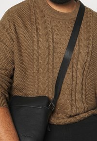 Hnědý svetr s kabelovým vzorem a vysokým límcem; malá černá kožená kabelka přes rameno s páskem spočívá na těle a ukazuje hladkou texturu.