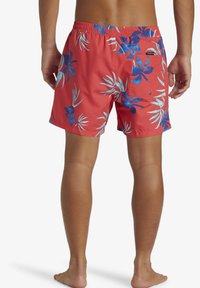 Red swim shorts with blue and white tropical floral pattern, elastic waistband, and back pocket with button closure, made of lightweight material.