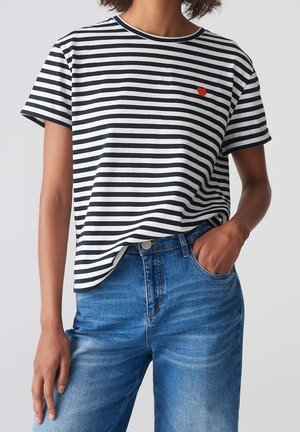 Femme portant un t-shirt à manches courtes rayé noir et blanc avec un détail cœur rouge, associé à un jean bleu taille haute, une main dans la poche.