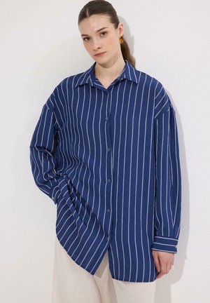 Vrouw draagt een loszittend blauw overhemd met witte verticale strepen en beige broek, staand tegen een witte achtergrond.