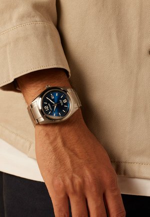 Stainless steel watch with a blue dial, silver hour markers, and a date display. Worn on a wrist with a textured beige sleeve in the background.