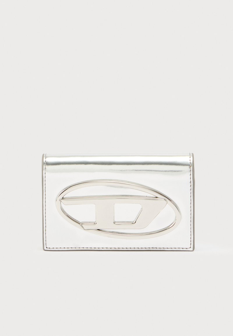Metalinis sidabro spalvos piniginė su lygiu paviršiumi, priekyje išsiskiria ovali sidabrinė detalė ir subtilūs siuvimo akcentai.