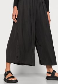 Sorte vide bukser lavet af letvægtsstoffer, med høj talje og et flowy design. Sammen med sorte sandaler med justerbare stropper.