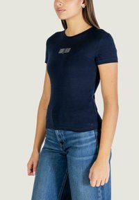 Tmavě modré přiléhavé tričko z jemné bavlny s krátkými rukávy. Má malý bílý logotyp na přední straně a kulatý výstřih.