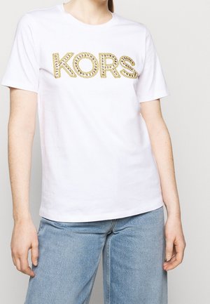 Personne portant une chemise blanche à manches courtes avec le texte "KORS" clouté en or et un jean en denim bleu clair, debout devant un fond uni.
