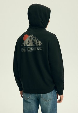 Mężczyzna w czarnej bluzie z kapturem z grafiką góry i czerwonego słońca oraz napisem "Henderson", w połączeniu z niebieskimi jeansami, stojący tyłem.