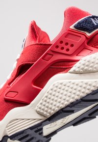 Röd idrottssko med en texturerad ovandel, gummisula och kontrasterande vita detaljer. Har anmärkningsvärda utskärningar och vadderade element.