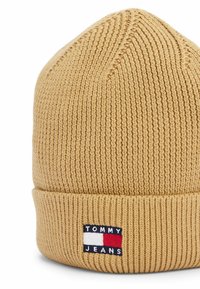 Tan brei-muts met een geribbelde structuur, voorzien van een omgeslagen rand en een patch met de tekst "TOMMY JEANS" in rode, witte en blauwe accenten.