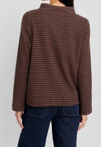 Žena oblečená v blůze s dlouhým rukávem, hnědo-červenými pruhy a tmavě modrými džíny, viděná ze zadu na pozadí světlé texturované zdi.