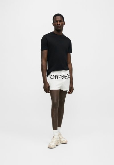 Muž stojaci v čiernom tričku s krátkym rukávom, bielych krátkych nohaviciach s nápisom "Off-White", bielych ponožkách a béžových teniskách na jednoduchom svetlom pozadí.