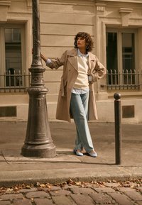 Cilvēks, kas valkā bēša krāsas lietusmēteļu un džinsus, stāv uz ielas, vienā rokā turot dekoratīvu laternu, netālu no bēša krāsas ēkas ar logiem.