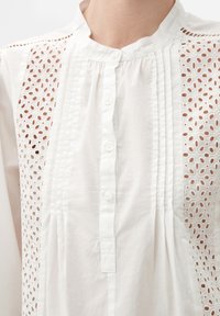 Blusa bianca con colletto a fascia, pattina con bottoni, pieghe verticali e pannelli in pizzo con motivi floreali e occhio di pernice sui lati anteriori.