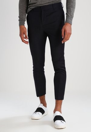 Mann trägt schwarze, gekürzte Slim-Fit-Hose, graues Langarmshirt und weiße Slip-On-Schuhe mit schwarzen Elastikbändern.