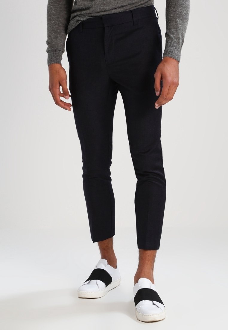 Homme portant un pantalon slim court noir, une chemise grise à manches longues et des chaussures blanches sans lacets avec des bandes élastiques noires.