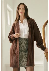 Femme debout sur un escalier, portant un cardigan marron en maille torsadée, une chemise blanche boutonnée et une jupe grise à motifs.