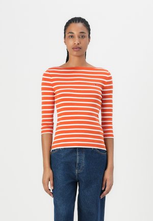 Orange og hvid stribet langærmet bluse med bådudskæring, parret med blå denim jeans. Subtilt logo på brystområdet.