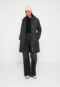 Czarny pikowany płaszcz z kapturem, z pionowym pikowaniem, zamkami w tonacji i zatrzaskami. Połączony z czarną bluzką i szerokimi dżinsami.