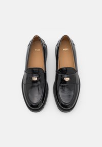 Mocassins en cuir noir à finition lisse, avec un logo doré sur le dessus et une semelle en caoutchouc antidérapante.