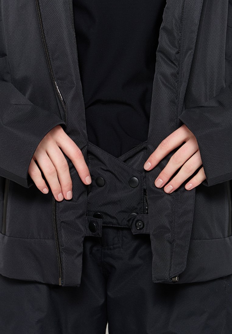 Mani che allacciano una giacca invernale nera con bottoni a pressione sopra una camicia e pantaloni neri.