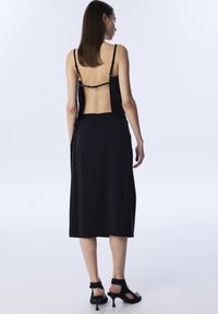 Vestido negro con tirantes finos, diseño de espalda abierta, tejido fluido y corte hasta los tobillos. Presenta una textura suave y un estilo minimalista.