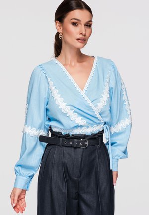 Kobieta nosząca jasnoniebieską bluzkę kopertową z białą koronkową lamówką oraz czarne plisowane spodnie z wysokim stanem i czarnym paskiem.