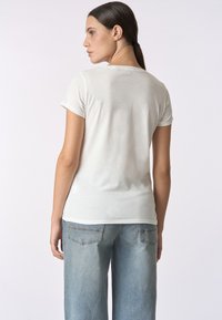 Maglietta bianca a maniche corte con collo tondo, texture liscia, design aderente e stile minimalista. Indossata con jeans a gamba larga di colore blu chiaro.