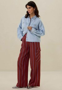 Hellblaues Denim-Hemd mit zwei vorderen Taschen, kombiniert mit weit geschnittenen, rostfarben gestreiften Hosen. Silberne spitze Schuhe vervollständigen das Outfit.