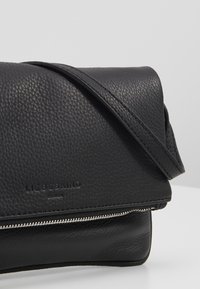 Černá texturovaná kožená crossbody taška s klopou a stříbrným zipovým detailem, s embosovaným názvem značky na předním panelu.