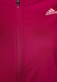 Giacca fucsia con zip, superficie liscia e testurizzata e piccolo logo bianco Adidas in alto a destra. La cerniera è verticale.