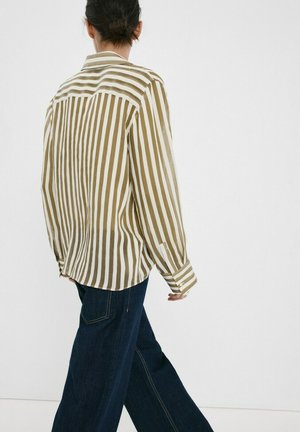 Persona con cabello oscuro recogido en un moño, vistiendo una camisa suelta de rayas verticales beige y blancas, y jeans oscuros de pierna ancha, caminando frente a un fondo blanco liso.