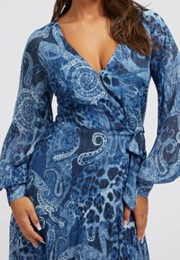 Robe enveloppante bleue présentant un motif paisley, des manches longues et une taille cintrée. Fabriquée en tissu léger et texturé.