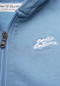 Blå zip-up hoodie gjord av mjukt tyg med en texturerad yta, med en vit broderad logotyp till vänster. Dragkedja i metallgrå.