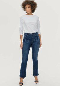 Jeans de mezclilla acampanados en azul oscuro con una cintura alta, combinados con una camisa blanca ajustada de manga larga y tacones negros destalonados.