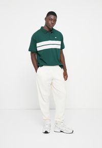 Grøn polo t-shirt med hvide vandrette striber, parret med cremefarvede joggingbukser og lysegrå sneakers. Ensfarvet baggrund.
