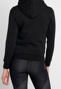 Persona che indossa un hoodie nero e leggings neri con texture, con un piccolo logo bianco sulla parte superiore della coscia, vista da dietro.