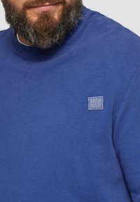 Blauw katoenen sweatshirt met een ronde hals en een klein, geborduurd logopatch op de linkerkant van de borst. De stof heeft een gladde textuur.